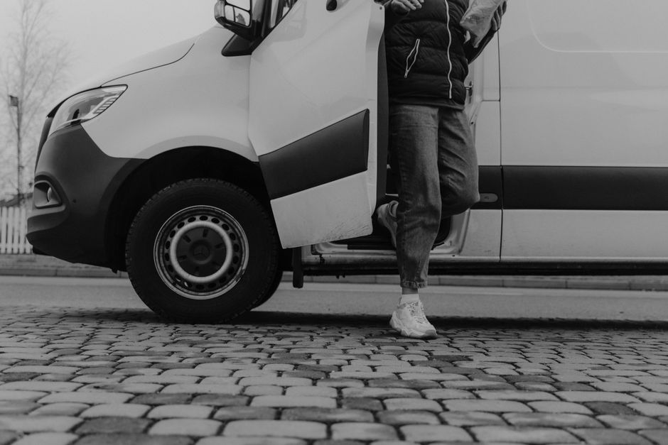 A man wearing a white t-shirt, jeans, and a black cap is seen carrying three cardboard boxes of varying sizes during a home relocation process outside a residential property. He is walking alongside a