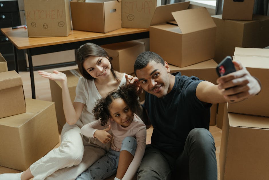 A young family sitting on a wooden-floored room surrounded by open cardboard boxes and packing materials, with a modern kitchen visible in the background. The mother, father, and their young daughter 