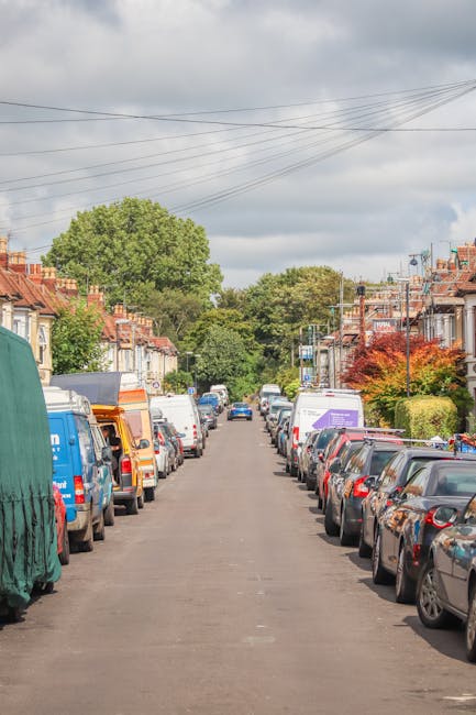 A residential street during daytime with rows of parked cars on both sides, including various models of sedans, hatchbacks, and vans, some covered with protective fabric. The street surface is asphalt, with a slight incline and no visible curb separation. In the background, there are trees with lush green foliage and well-maintained small front gardens with bushes and colorful plants. Overhead, there are power lines stretching across the sky, which is partly cloudy with grey clouds. The scene is quiet with no pedestrians or moving vehicles visible. This setting provides a typical environment for house removals and furniture transport services offered by manwithvanwalthamstow.co.uk, suitable for discussions about home relocation logistics in narrow streets like those in Walthamstow.