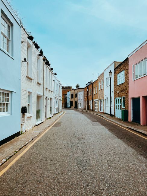 A narrow residential street in Walthamstow with a gentle curve, featuring a row of terraced houses painted in pastel shades including light blue, white, beige, and pink. The houses have brickwork, large windows, and some with small front gardens or planters by the doors. A string of bulbs is strung across the top of the buildings, extending along the street. The asphalt road is flanked by a narrow pavement on each side, with a few wheelie bins visible near the house entrances. The scene is well-lit with natural daylight, highlighting the clean, quiet atmosphere typical of a house removal environment in Walthamstow. The image may relate to a house relocation or furniture transport process, as it shows an accessible street suitable for van parking and loading activities, with no vehicles present in the immediate view. This setting aligns with the services offered by man with van Walthamstow for home removals across narrow streets like those in Walthamstow Village.